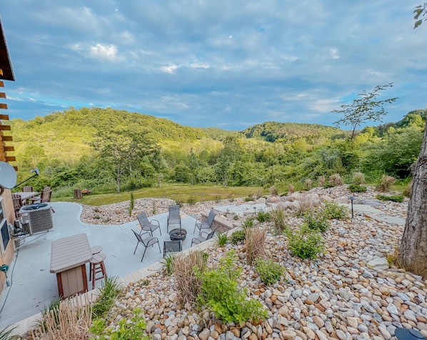 An outdoor patio with a seating area, surrounded by a garden with various plants and a view of a forested hillside. - Ash Ridge Lodge in Hocking Hills (Laurelville)