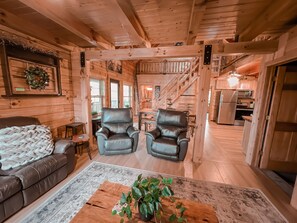 A cozy cabin interior with a living area featuring a sofa, two leather chairs, a coffee table with a plant, and a staircase leading to the kitchen.