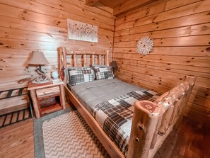 A wooden cabin bedroom with a bed, nightstands, a lamp, a wall hanging, and a decorative wall piece. - Ash Ridge Lodge in Hocking Hills (Laurelville)