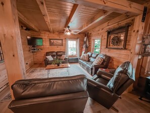 A cozy log cabin interior with leather sofas, a wooden coffee table, and a ceiling fan.