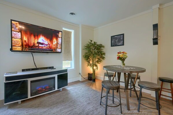 A living room with a flat-screen TV, a fireplace, a dining table with chairs, a potted plant, and a vase with flowers.