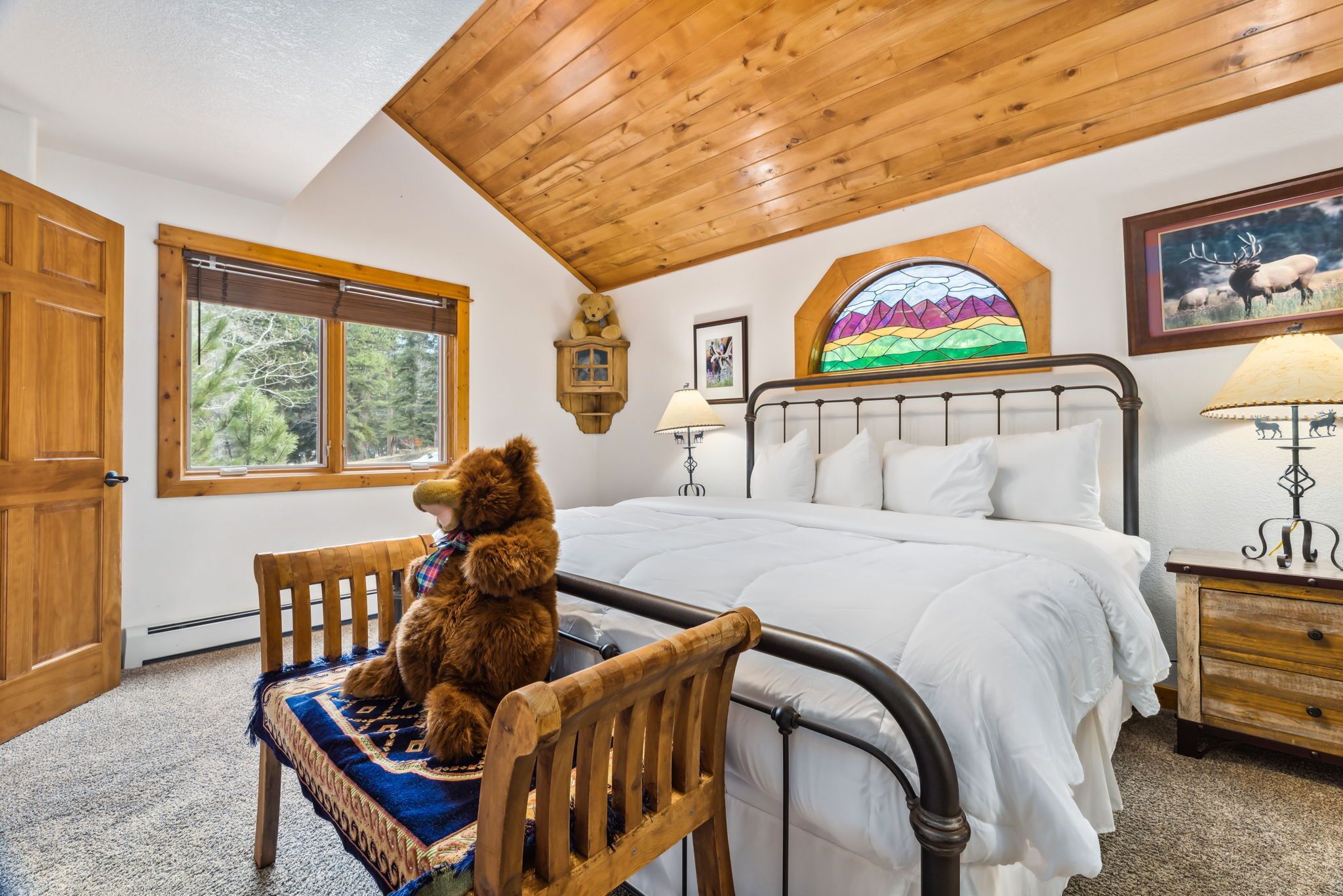 Un dormitorio con techo de madera, una cama con ropa de cama blanca, una silla de madera con un osito de peluche, una ventana con vistas a árboles y un vitral.