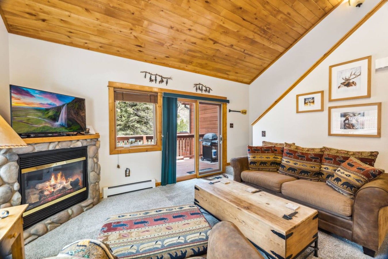 A cozy living room with a stone fireplace, a television, a sofa with patterned cushions, and a wooden ceiling.