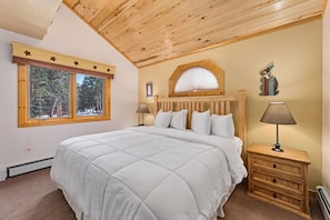 A bedroom with a large bed, a window with a view of trees, a wooden headboard, and a nightstand with a lamp.
