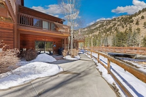 A wooden cabin with a balcony, a concrete pathway, and a wooden bridge over a stream.