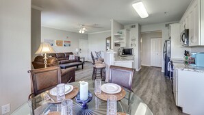 A modern kitchen with white cabinets, a microwave, and a refrigerator. There is a dining area with a glass table and chairs, and a living room with a couch and a coffee table.