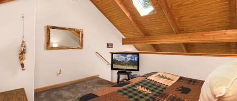 A bedroom with a sloped ceiling, a wooden bed frame, a quilt, a television, and a small window.