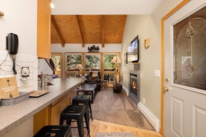 A kitchen with a bar area, wooden ceiling beams, a fireplace, and a view of trees through large windows. - Scenic Riverfront Escape • RMNP • River Views (Estes Park)