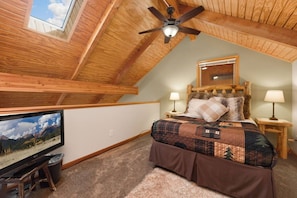 A bedroom with a sloped ceiling, a bed with a quilt, two bedside lamps, a wooden headboard, a television, and a window with a mountain view. - Scenic Riverfront Escape • RMNP • River Views (Estes Park)