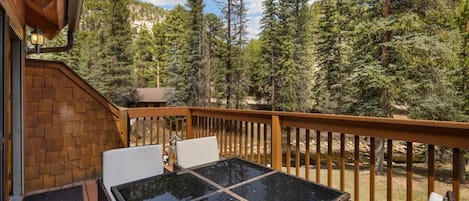 A wooden deck with a glass table and chairs, surrounded by trees.