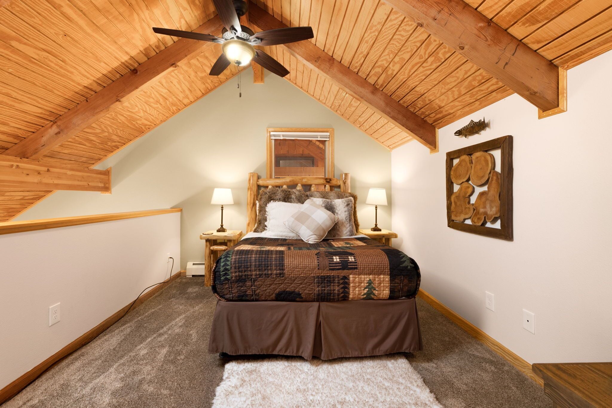 Quarto com teto inclinado, cama com colcha estampada, duas luminárias de cabeceira, ventilador de teto e um quadro na parede.