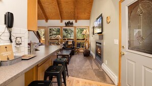 Una cocina con techo de madera, una zona de bar con taburetes, una chimenea y un ventanal con vistas a árboles.
