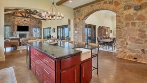 A spacious kitchen with a large island, stone walls, and a dining area with a stone archway. - Luxe Tuscan Villa on 1 Acre with Pool & Pickleball, Sleeps 18, 9 mins to DT (Sunset Valley)