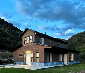 Una casa moderna de dos pisos con un amplio porche delantero, un patio cubierto y vistas a un paisaje montañoso.