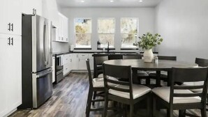 Una cocina moderna con armarios blancos, un refrigerador de acero inoxidable, una mesa de comedor con sillas y ventanas amplias con vistas a árboles.