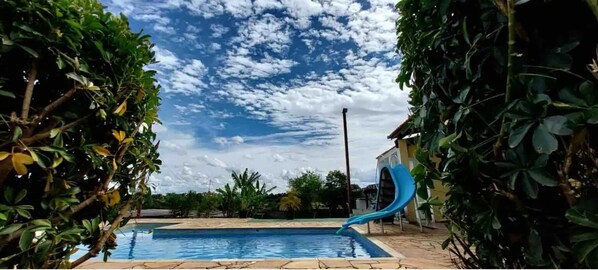 A swimming pool with a blue slide, surrounded by lush greenery and a clear sky. - Chácara Roteiro do Vinho com Wi-fi (Canguera)