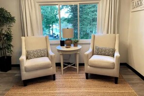 Two white armchairs with patterned cushions, a small round table with a lamp, and a view of trees through a window.