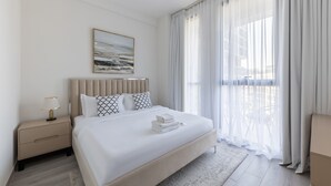 A modern hotel room with a large bed, a bedside table, a lamp, and a view of the outdoors through sheer curtains.