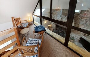A cozy balcony with wooden chairs, a small table, and a glass railing overlooking a stone fireplace. - Fermette Bretonne Typique en Pierres au Bout du Bout du Finistère Pays des Abers (Plouguerneau)