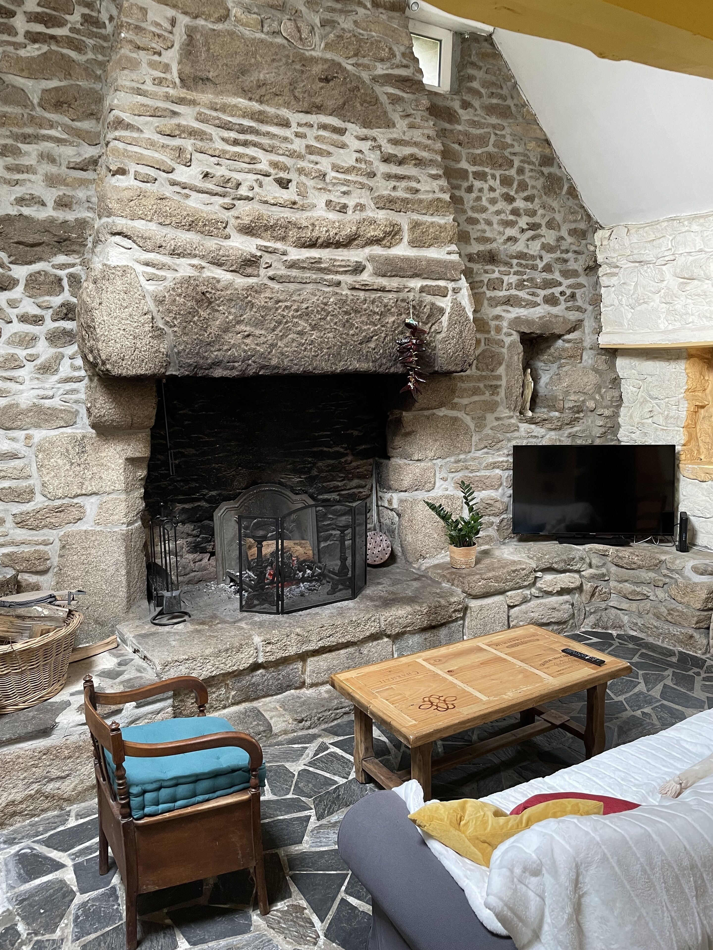 A stone fireplace with a television mounted above it, a wooden table, a cushioned chair, and a sofa with a throw pillow.