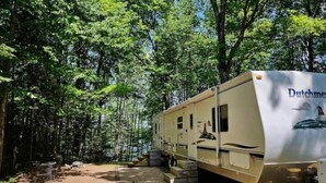 Un veicolo ricreativo (RV) parcheggiato in una zona boschiva con terreno sterrato e una piccola veranda in legno.