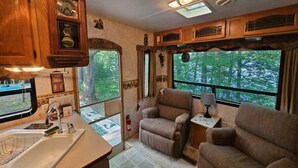 Un intérieur de camping-car compact comprenant un coin cuisine, un espace salon et de grandes fenêtres offrant une vue sur la forêt.