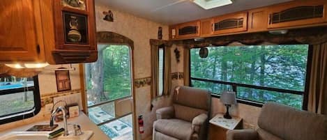 Un intérieur de camping-car compact comprenant un coin cuisine, un espace salon et de grandes fenêtres offrant une vue sur la forêt.