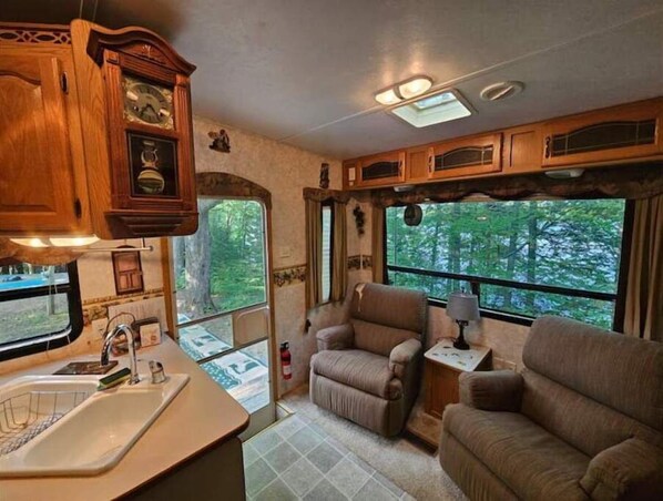A compact RV interior with a kitchenette, seating area, and large windows offering a view of the forest.