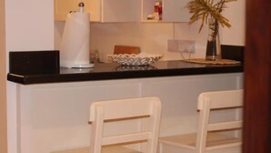 Una cocina con encimera negra, dos sillas blancas, un lavabo blanco y un florero con planta.