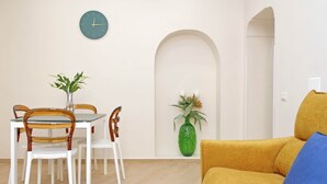 Ein Essbereich mit einem weißen Tisch und Stühlen, einer grünen Vase mit Pflanzen, einer Uhr an der Wand und einem gelben Sofa mit einem blauen Kissen.