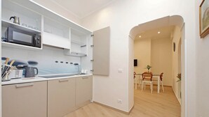 Eine moderne Küche mit weißen Schränken, einer Mikrowelle und einer Kaffeemaschine. Ein Essbereich mit Tisch und Stühlen ist durch einen offenen Durchgang sichtbar.