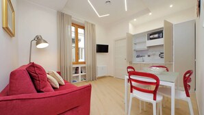 A modern living room with a red sofa, white dining table, and red chairs. There is a TV mounted on the wall and a window with curtains. - 4BNB - Ottaviano Vatican Flat - Red (Rome)