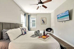 Ein Schlafzimmer mit einem Bett, einem Deckenventilator, einem Fenster mit Jalousien, einem Gemälde und einem an der Wand befestigten Fernseher.