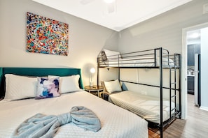 Hotellihuone, jossa on kerrossänky, yksi erillinen sänky, värikäs abstrakti maalaus, yöpöytälamppu ja näkymä keittiöön.