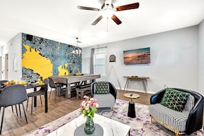 Ein modernes Wohnzimmer mit Essbereich, Deckenventilator, einer Wand mit Stadtbild-Mural und einem Fernseher, der eine Sonnenuntergangsszene zeigt.