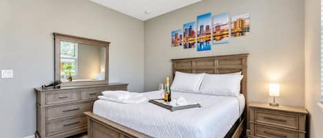 Quarto com cama grande, cômoda, mesa de cabeceira, ventilador de teto e uma pintura com vista para a cidade.