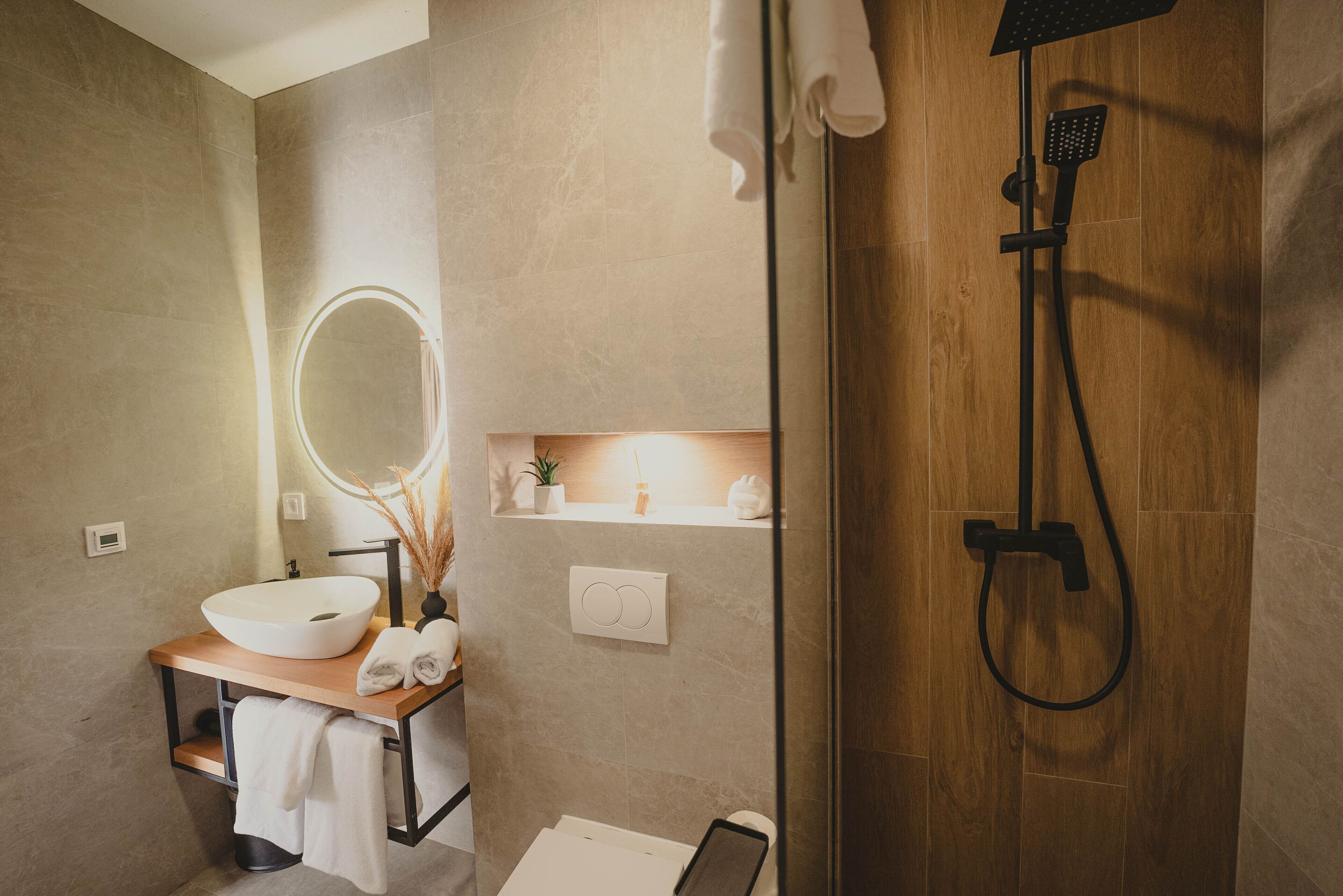 Een moderne badkamer met een houten douche, een ronde spiegel, een witte wastafel en een plank met toiletartikelen.