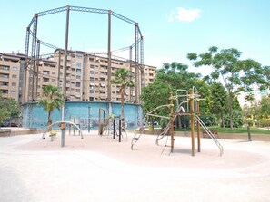 A playground with swings and climbing structures, surrounded by a blue wall and palm trees, with a multi-story building in the background. - Port Area II. WiFi + Air Condition (Valencia)