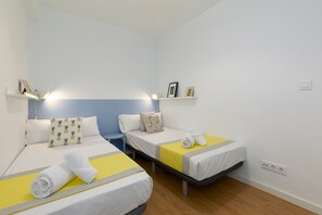 A hotel room with two single beds, each with a yellow and white bedspread, a white pillow, and a rolled white towel. There is a small blue chair and a shelf with framed pictures. - Port Area II. WiFi + Air Condition (Valencia)