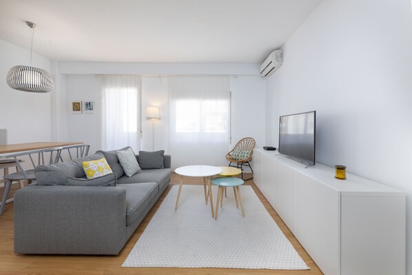 Ein modernes Wohnzimmer mit grauem Sofa, einem kleinen runden Tisch, einem Fernseher und einem Essbereich mit Tisch und Stühlen.