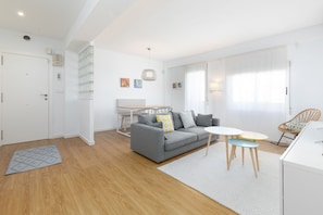 Ein modernes Wohnzimmer mit einem grauen Sofa, einem kleinen runden Tisch und einem Korbstuhl. Eine weiße Tür und ein Fenster mit Vorhängen sind ebenfalls zu sehen.