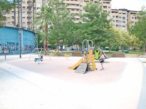 A playground with various play equipment, surrounded by trees and a building in the background.