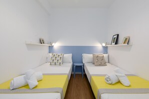 A room with two beds, each with a yellow bedspread and white pillows. There is a small blue stool in the center, and a shelf above the beds holding framed pictures. - Port Area II. WiFi + Air Condition (Valencia)