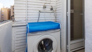 Eine kompakte, weiße, wandhängende Waschmaschine mit blauem Deckel, neben einer Schiebeglastür, an einer weißen Außenwand mit sichtbaren Leitungen.