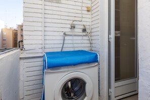 A front-loading washing machine with a blue cover, mounted on a white exterior wall with visible plumbing. - Port Area II. WiFi + Air Condition (Valencia)