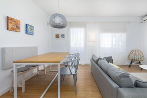 Ein modernes Wohnzimmer mit einem Holzesstisch, grauen Stühlen, einem grauen Sofa und einem Korbstuhl. An den Wänden hängen zwei Gemälde, eine Pendelleuchte und ein Fenster mit Vorhängen.