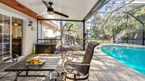 Un patio al aire libre con piscina, ventilador de techo, una silla con cojines y una mesa con fruta.