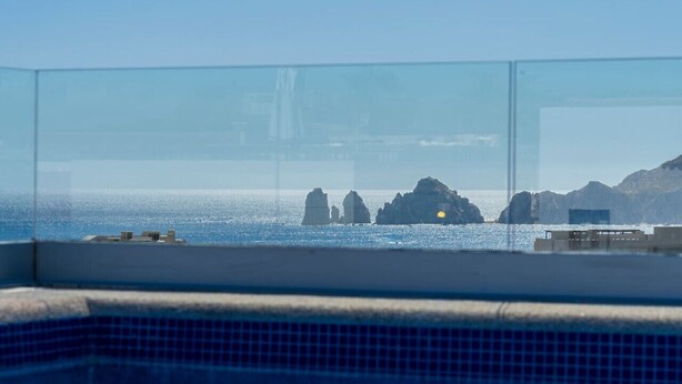 Klarer Blick auf das Meer mit Felsformationen, ein Gebäude in der Ferne und ein Poolbereich mit Fliesenboden.