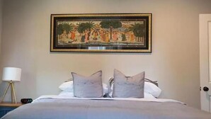 Una cama bien hecha con una colcha gris y dos almohadas, un cuadro enmarcado encima, una lámpara de noche y una puerta a la derecha.