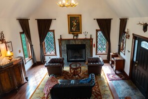 Ein stilvolles Wohnzimmer im traditionellen Stil mit Kamin, Ledersofas, einer Kristallleuchtre und einem großen Fenster mit Blick auf Grünflächen.